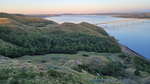A scenic view of rolling hills and a river, with a bridge in the distance under a clear sky at sunset.