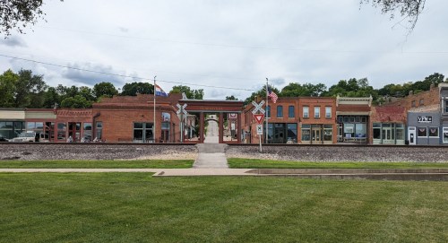 Historic buildings line a street in a small town, featuring flags and a grassy area in the foreground.
