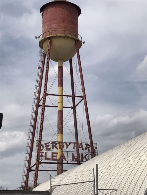 A tall, red water tower with a sign reading "Derby Park Flea Market" against a cloudy sky.