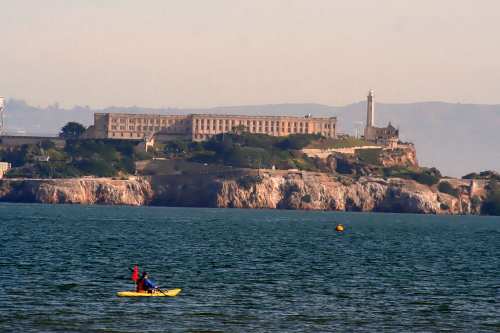 A kayaker paddles in the water near Alcatraz Island, featuring its historic buildings and lighthouse in the background.