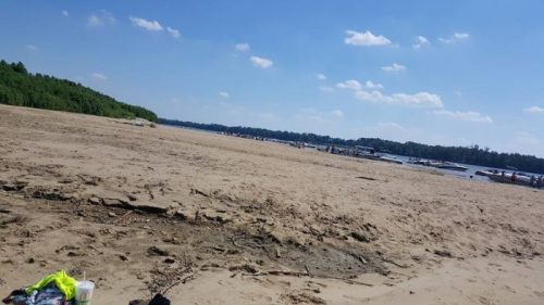 A sandy beach under a clear blue sky, with boats in the distance and greenery along the shore.
