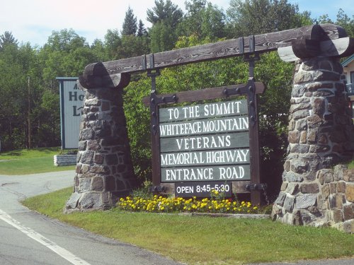 Sign for Whiteface Mountain Veterans Memorial Highway, featuring stone pillars and flowers, with operating hours listed.