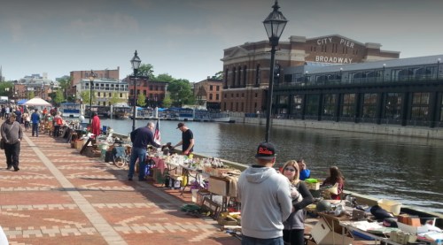 A bustling waterfront market with vendors and shoppers along a brick pathway by the water, surrounded by buildings.