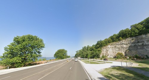 A scenic view of a highway alongside a river, with lush trees and a rocky cliff on one side under a clear blue sky.