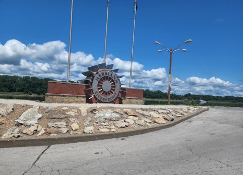 Sign for Hermann Riverfront with flags and a scenic blue sky in the background. Rocky area in the foreground.