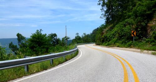 A winding road curves through lush greenery under a clear blue sky, with a caution sign for a sharp turn ahead.