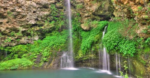 A serene waterfall cascading into a lush green pool, surrounded by rocky cliffs and vibrant foliage.