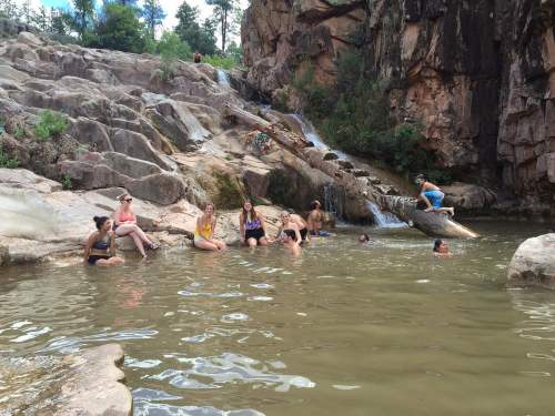 A group of people relaxes in a natural pool surrounded by rocks and a waterfall in a scenic outdoor setting.