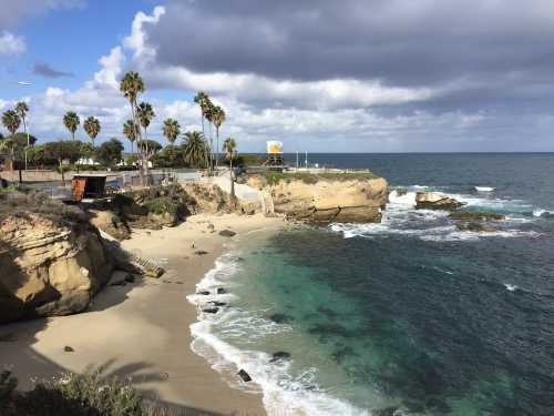 Coastal view featuring sandy beach, rocky shore, palm trees, and waves under a partly cloudy sky.