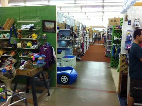 A wide aisle in a thrift store filled with shelves of various items, including toys, clothes, and home goods.