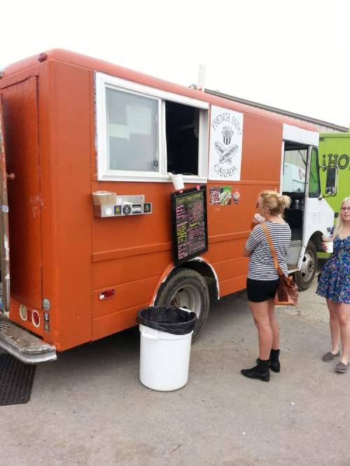 A vibrant orange food truck with a menu board, while a woman in a striped shirt waits nearby.