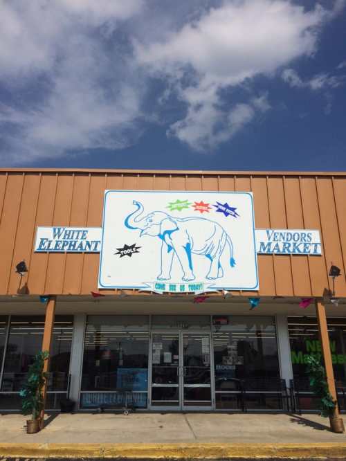 Sign for "White Elephant Vendors Market" featuring an elephant graphic, with colorful stars and a blue sky backdrop.