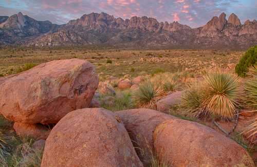 Rocky landscape with large boulders in the foreground and a mountain range under a colorful sky at sunset.