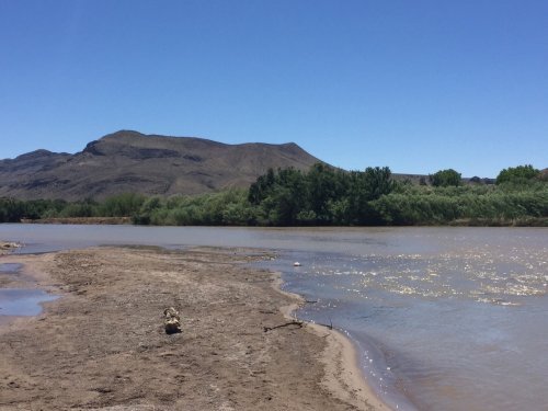 A serene riverbank scene with a sandy shore, gentle water, and mountains in the background under a clear blue sky.
