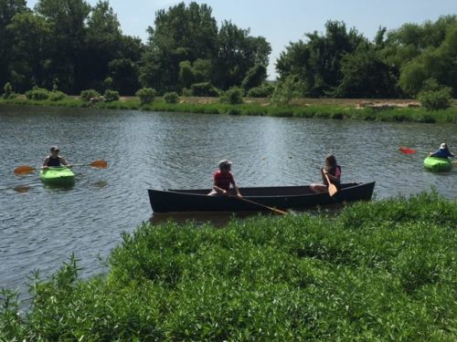 Two kayakers paddle on a calm river while a person sits in a canoe, surrounded by lush greenery.