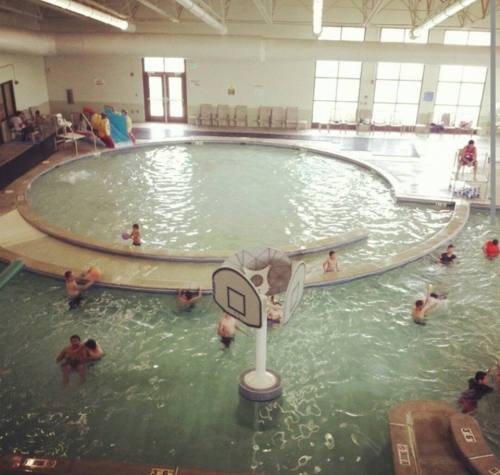 A spacious indoor pool with children playing, a basketball hoop, and lounge chairs around the edge.
