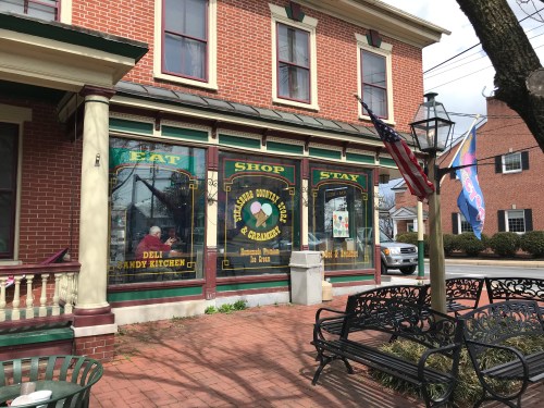 A brick building with colorful shop windows displaying "Eat," "Shop," and "Stay," featuring a deli and candy kitchen.