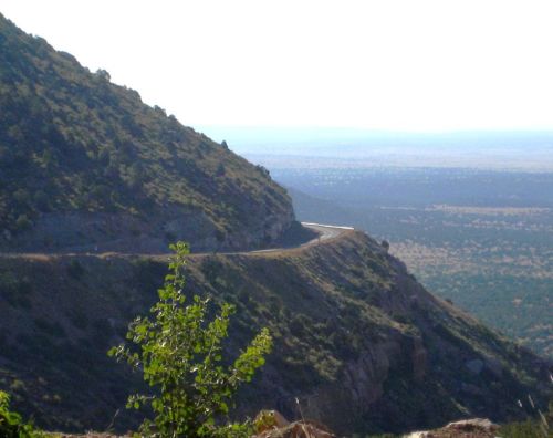 A winding road curves along a mountainous landscape, with a vast view of the plains in the distance.