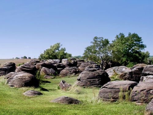 A landscape featuring large, rounded rocks scattered across green grass, with trees and a clear blue sky in the background.