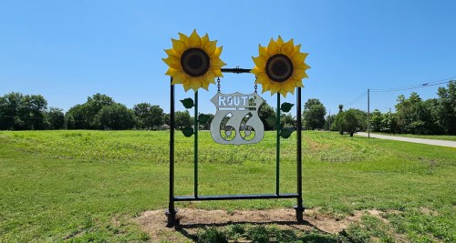 Sign for Route 66 adorned with two large sunflowers, set against a clear blue sky and green landscape.