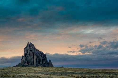 A dramatic rock formation rises from a grassy plain under a colorful, cloudy sky at sunset.