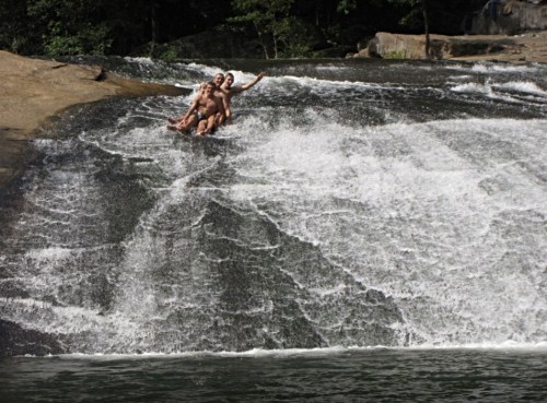 Three people slide down a rocky waterfall, splashing water around them in a natural setting.
