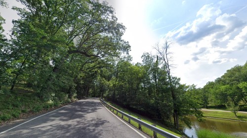 A winding road surrounded by lush green trees, with a calm body of water on one side under a partly cloudy sky.