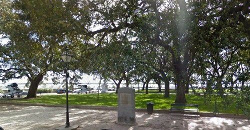 A park scene with large trees, a stone monument, a bench, and a view of the water in the background.