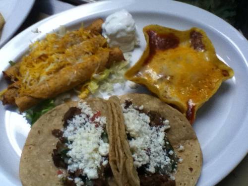 A plate of Mexican food featuring tacos, taquitos, cheese, sour cream, and a baked dish with melted cheese.