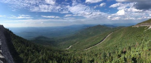 Panoramic view of lush green mountains under a blue sky with scattered clouds, showcasing winding roads and distant peaks.
