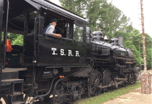 A vintage steam locomotive with "T.S.R.R." on the side, parked beside a signal post in a wooded area.