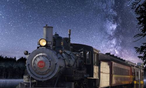 A vintage steam train parked under a starry night sky, with the Milky Way visible above.