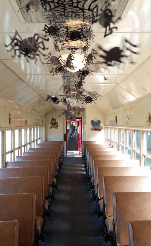 Interior of a train car decorated with hanging spiders, featuring rows of brown seats and a view of the exit.