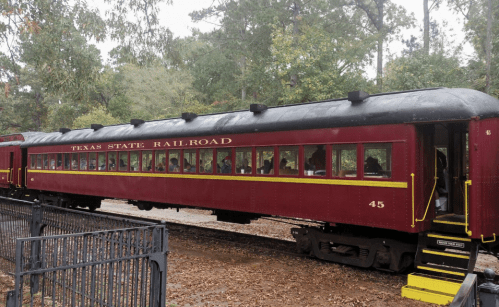 A red vintage train car labeled "Texas State Railroad" parked among trees with passengers visible inside.