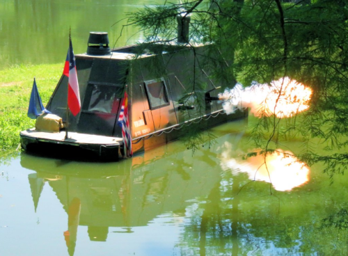 A black boat on a calm water surface, with flags and a plume of smoke rising from the rear.