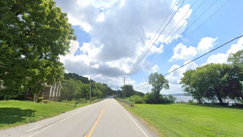 A scenic road lined with trees, leading towards a distant hill under a partly cloudy sky.