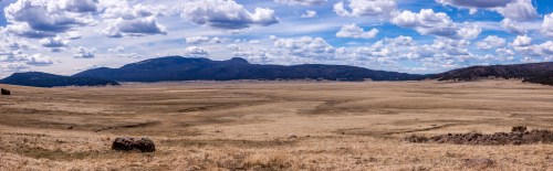 A panoramic view of a vast, dry landscape with rolling hills and a blue sky filled with fluffy clouds.