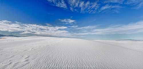 A vast white sand dune landscape under a bright blue sky with scattered clouds.