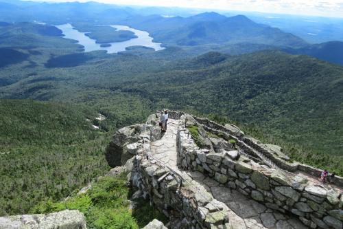 A scenic view from a mountain summit, showcasing lush green forests and a winding lake in the distance under a blue sky.