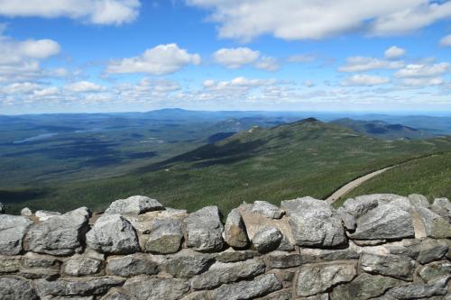 A scenic view of rolling green mountains under a blue sky with fluffy clouds, framed by a stone wall in the foreground.
