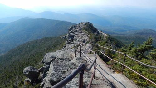 A rocky mountain trail with a railing, overlooking a vast landscape of mountains and valleys in the distance.