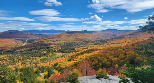 A vibrant autumn landscape with colorful trees, rolling hills, and a clear blue sky dotted with clouds.