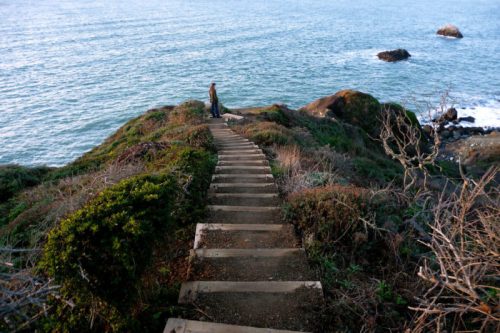 A winding staircase leads down a rocky hillside to the ocean, with a person standing near the water's edge.