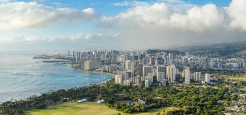 Aerial view of a coastal city with skyscrapers, lush greenery, and a blue ocean under a partly cloudy sky.