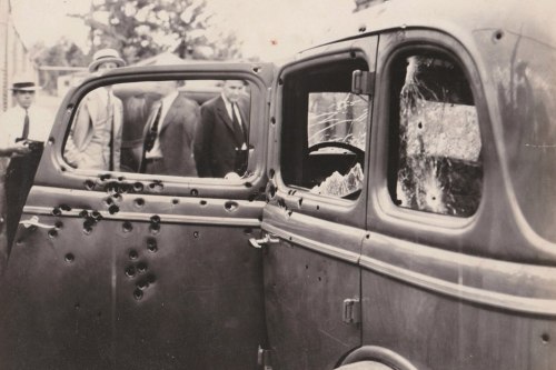 A vintage car with bullet holes in the door and window, with people observing in the background.