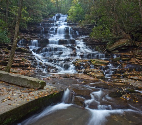 A serene waterfall cascading down rocky steps, surrounded by lush greenery and autumn leaves.
