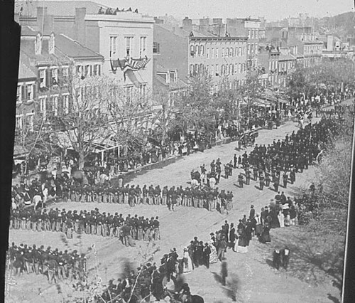 A historic black-and-white photo of a large crowd gathered on a street, with people in uniform and buildings in the background.