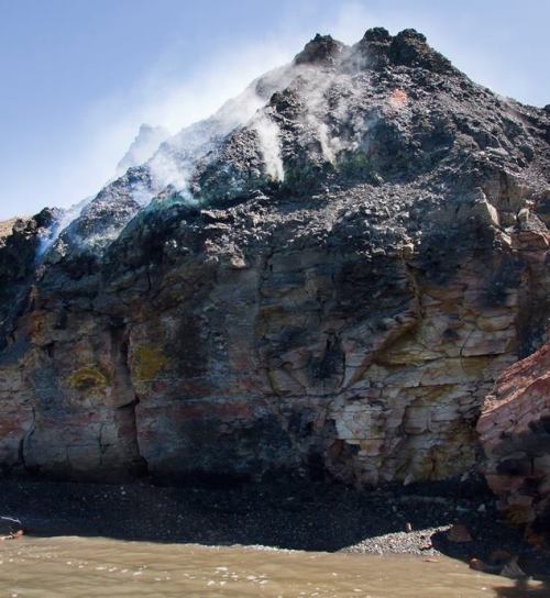 A rocky volcanic landscape with smoke rising from the summit, near a body of water.