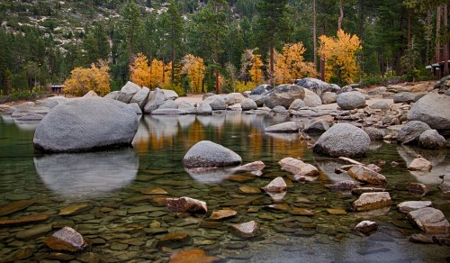 A serene lake surrounded by autumn trees, with large rocks and clear water reflecting the landscape.