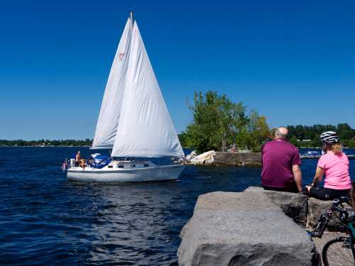 A sailboat glides on calm water while two people sit on a stone pier, enjoying the sunny day.